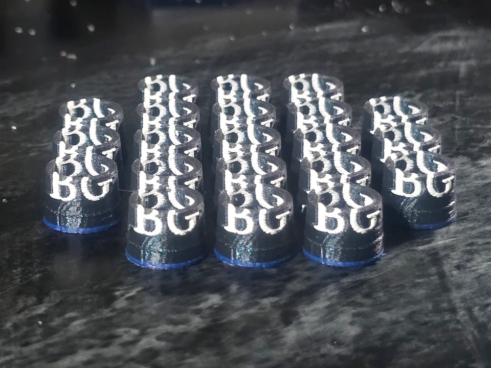 A batch of black 3D-printed ferrules with white BG initials and a blue base ring, fresh off the printer